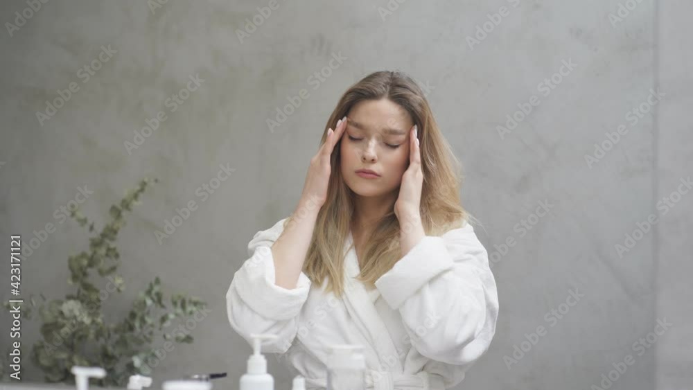 Young beautiful woman is worried about facial skin condition, looking at mirror in bathroom, in the morning, wearing bathrobe. Daily hygiene and skincare. Beauty concept Young beautiful woman is worried about facial skin condition, looking at mirror in bathroom, in the morning, wearing bathrobe. Daily hygiene and skincare. Beauty concept