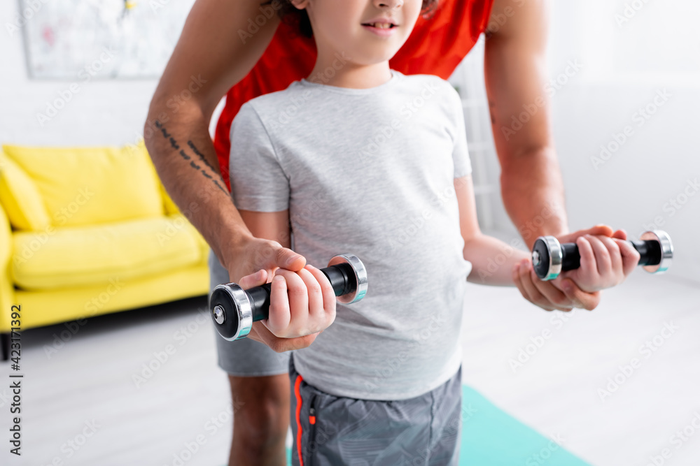Cropped view of father helping son with dumbbells during work out at home Cropped view of father helping son with dumbbells during work out at home