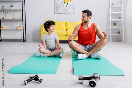 Smiling arabian father and son sitting in yoga pose near dumbbells at home