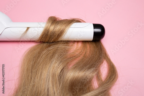 Curling blonde hair on a large diameter curling iron on a pink background. Curl care, hair styling.