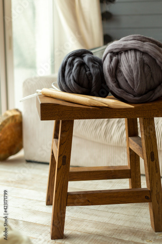Two large balls of merino wool lying on a wooden stool with wooden knitting needles against the background of a window and a white sofa © riakhinantonUkraine