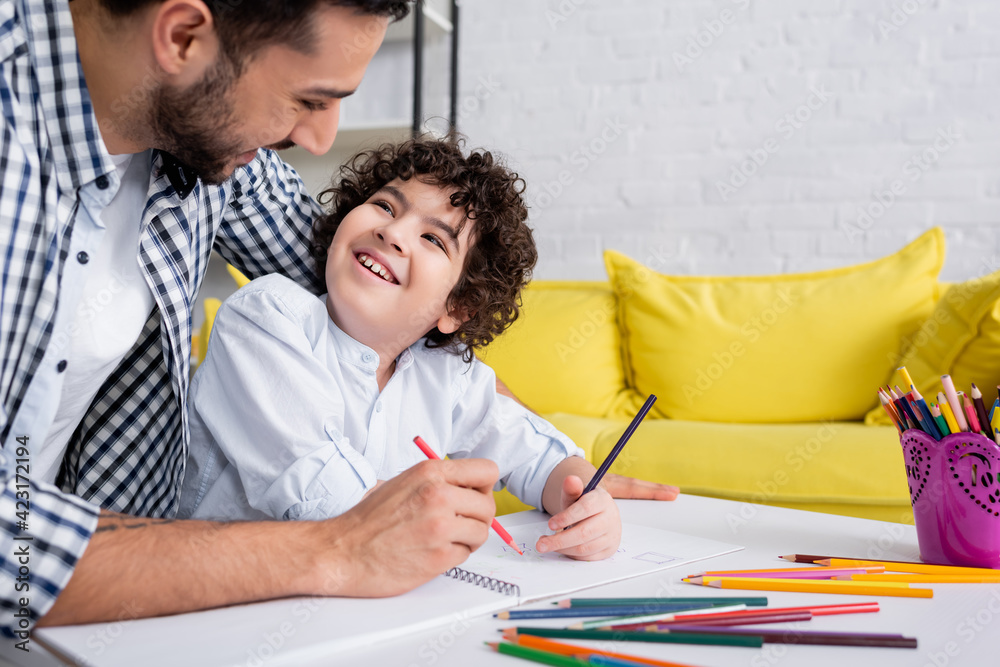 joyful muslim kid and father looking at each other while drawing with pencils together joyful muslim kid and father looking at each other while drawing with pencils together