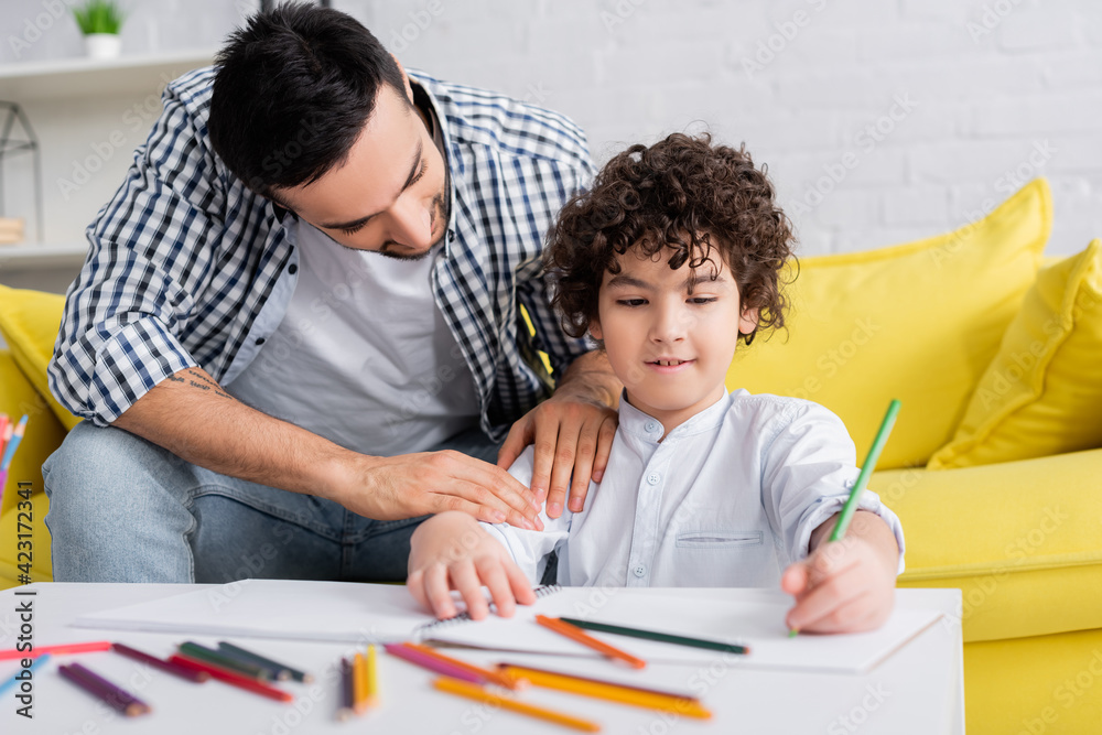 Naklejka premium young arabian man touching son drawing with pencil in sketchbook on blurred foreground