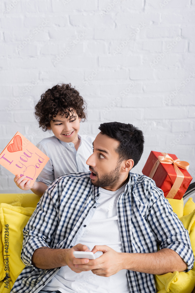 Φωτογραφία Surprised Muslim man near cheerful son holding I love you dad card and gift box