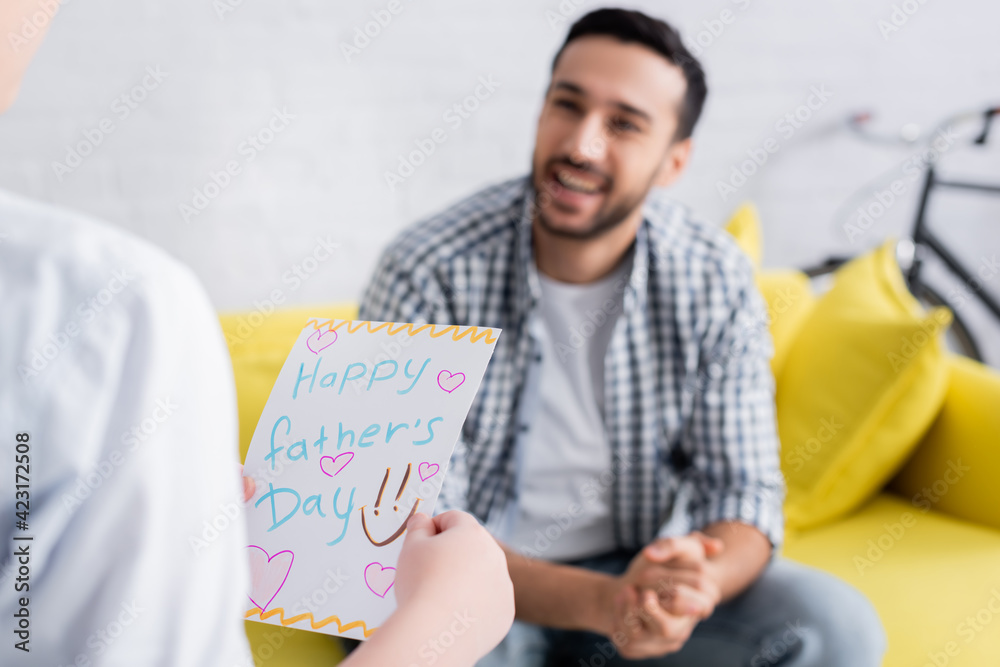 Cheerful Arabian man looking at son holding happy fathers day card on blurred foreground Cheerful Arabian man looking at son holding happy fathers day card on blurred foreground
