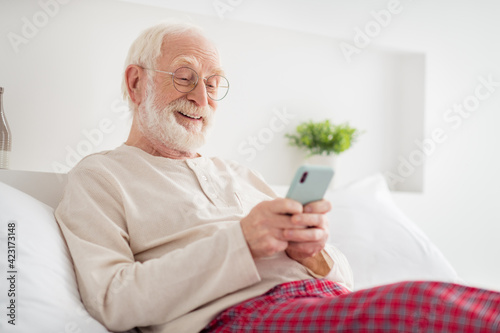 Photo of aged man pensioner happy positive smile sit bed home chat type sms look read browse cellphone feel young