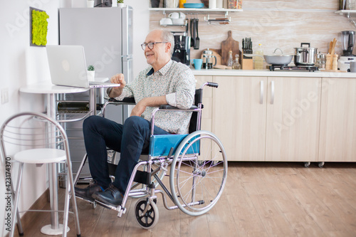 Senior man in wheelchair using laptop in the kitchen. Disabled senior man in wheelchair having a video conference on laptop in kitchen. Paralyzed old man and his wife having a online conference.