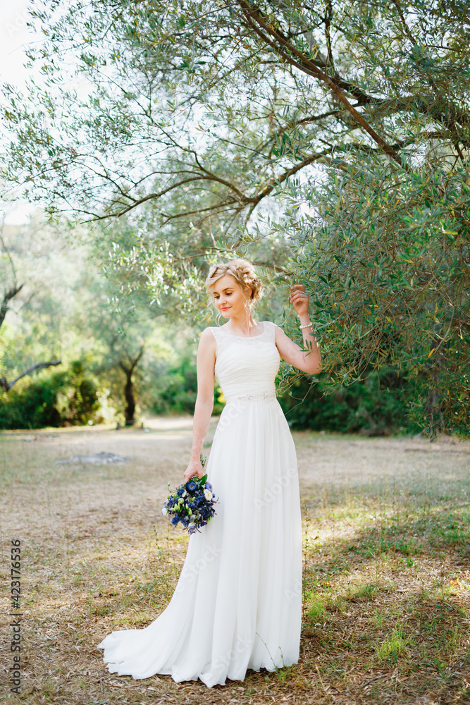 A tender bride with a bouquet of blue flowers stands by an olive tree and touches the branches with her hand 