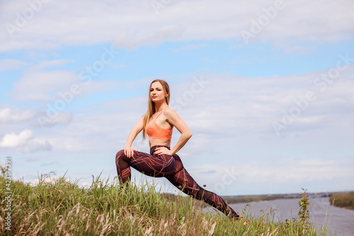 Woman is stretching in lunge outdoors on the river bank in front of the blue sky