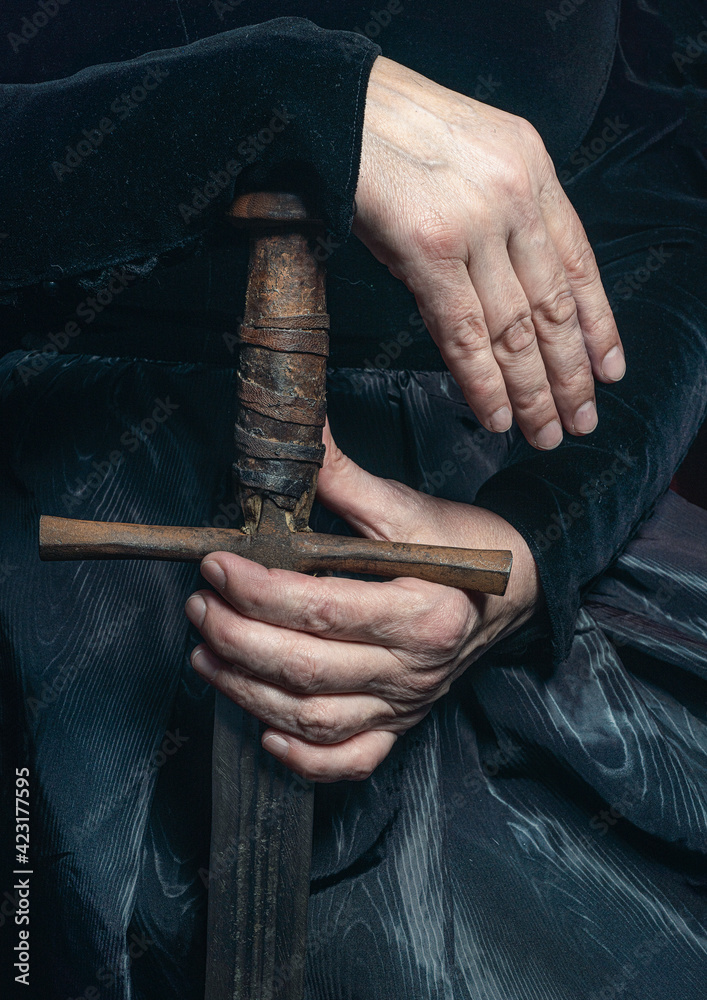 Female hands holding a sword, cropped photo. Fabulous creature, historical character, fantasy queen, witch, in a fairy costume. Woman, model after 60, in the studio, dark background
