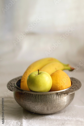 Antique bowl with bananas, apples and lemons. Selective focus, white textile background.