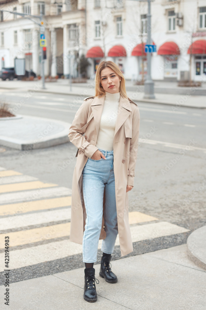 Young beautiful blonde hair woman in fashion clothes: beige trench coat, black boots and jeans crossing the road street. City lifestyle portrait.