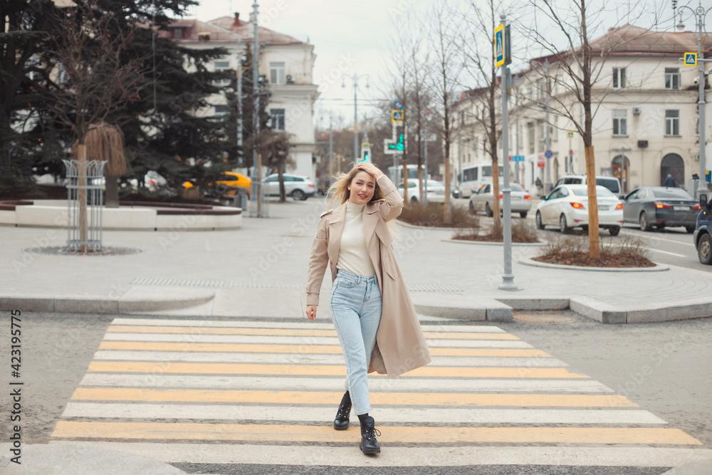 Young beautiful blonde hair woman in fashion clothes: beige trench coat, black boots and jeans crossing the road street. City lifestyle portrait.