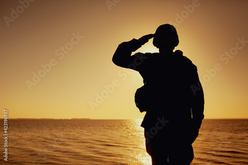 Silhouette of soldier in combat helmet and ammunition saluting on background of sunset sky. Army special forces fighter, Marines rifleman showing respect, greeting officer with salute gesture