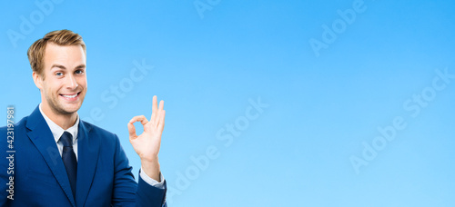 Portrait of confident businessman showing ok hand sign gesture, on blue background. Business success concept. Young happy smiling man at studio.