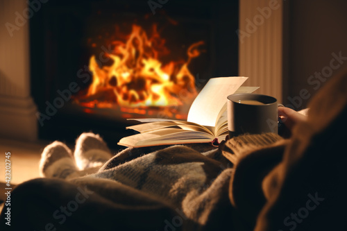 Woman with cup of drink and book near fireplace at home, closeup photo