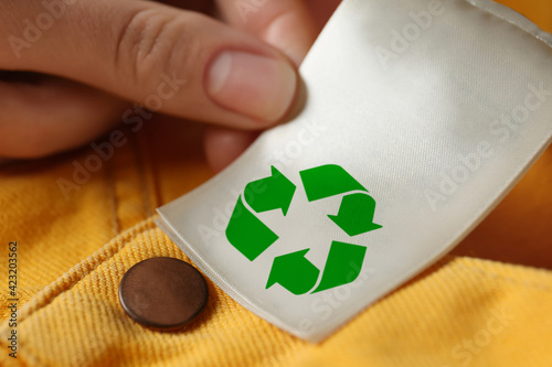 Woman showing clothing label with recycling symbol on yellow jeans, closeup photo