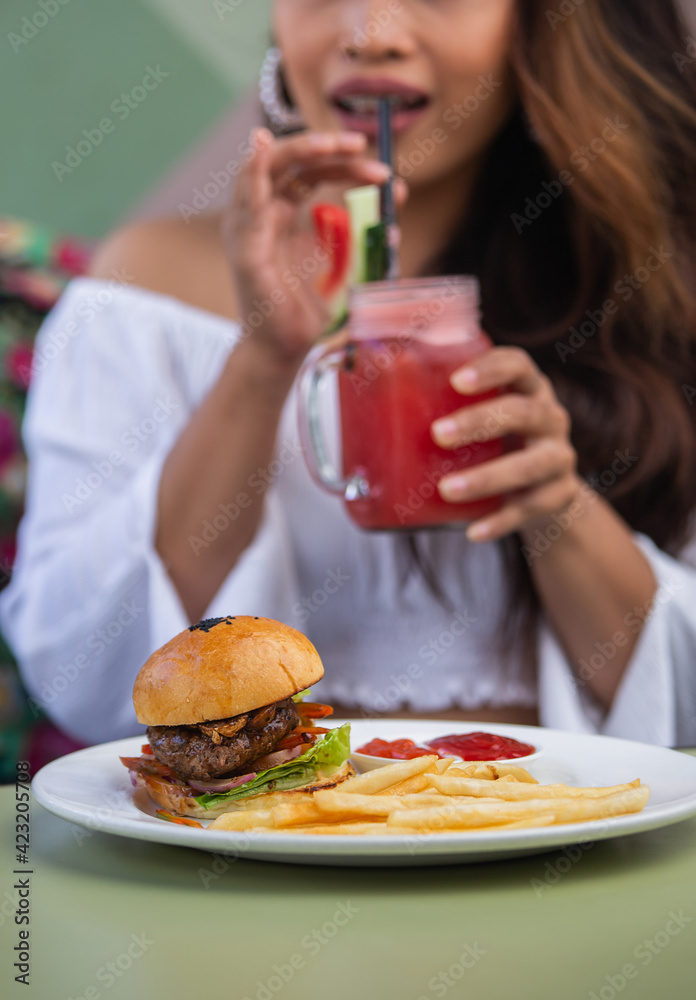 Close up of young woman eating unhealthy food in cafe or restaurant: burger, french fries potatoes and smoothie