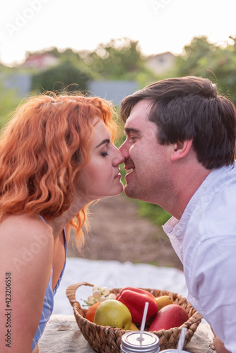 Young red-haired woman kisses a dark-haired man at breakfast in the morning. A couple in love traveling out of town on a weekend in nature in their trailer truck. Fruits lie on a wooden table.