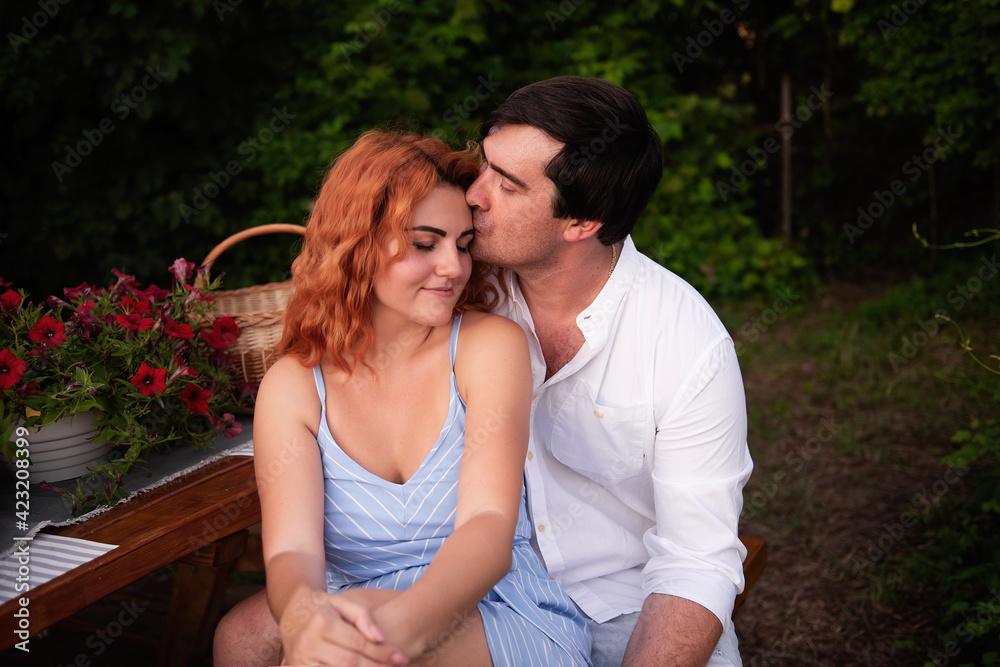 Fototapeta premium A young brunette man gently kisses a beautiful red-haired girl on the forehead, she laughs. Close-up portrait of couple in love, resting on picnic at trailer truck. Lovers kiss, hug. Travel honeymoon