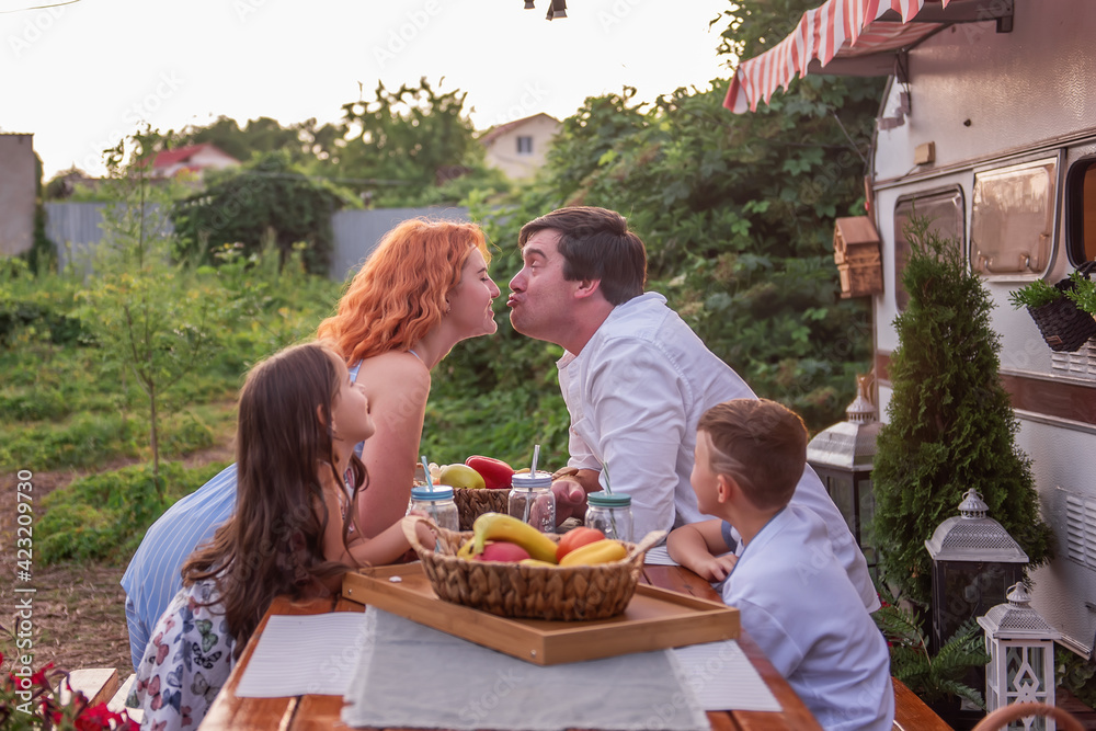 Happy family sits at a wooden table, drinks lemonade together by a trailer truck. Picnic, weekend camping with children. Travel in a motor home. Parents kiss, kids watch, laugh. Rest outside the city