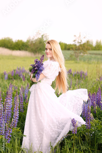 Young attractive long-haired blonde in a white vintage dress with a bouquet of lupins among a purple field. The concept of nature and romance.