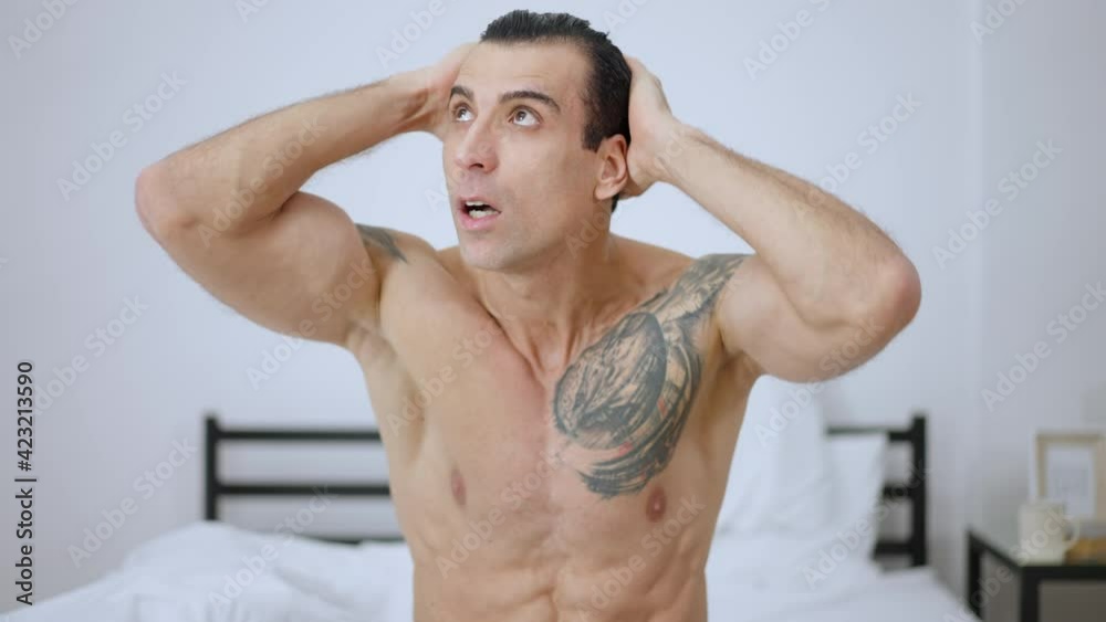 Middle shot of angry stressed man sitting on bed thinking. Unhappy Middle Eastern tattooed guy in bedroom at home in the morning. Stress and anger concept. Middle shot of angry stressed man sitting on bed thinking. Unhappy Middle Eastern tattooed guy in bedroom at home in the morning. Stress and anger concept.