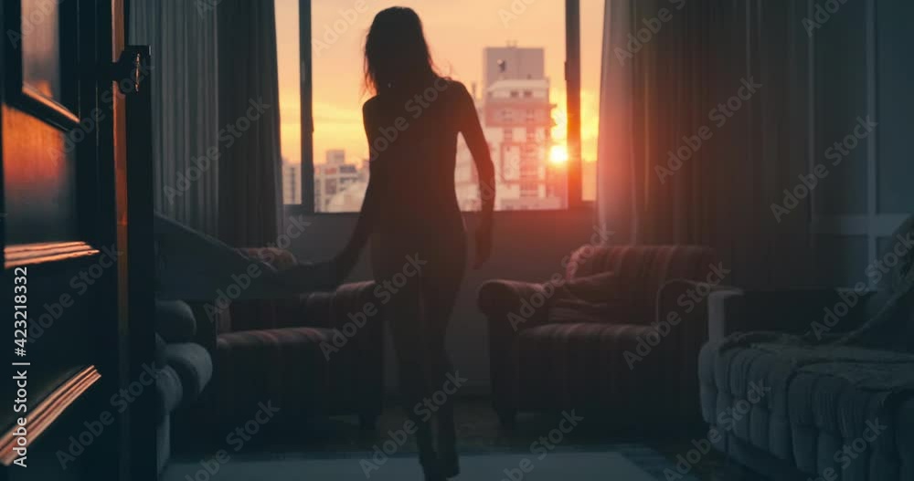 Young woman takes off her summer dress and dances near the wide window in the apartment at sunset