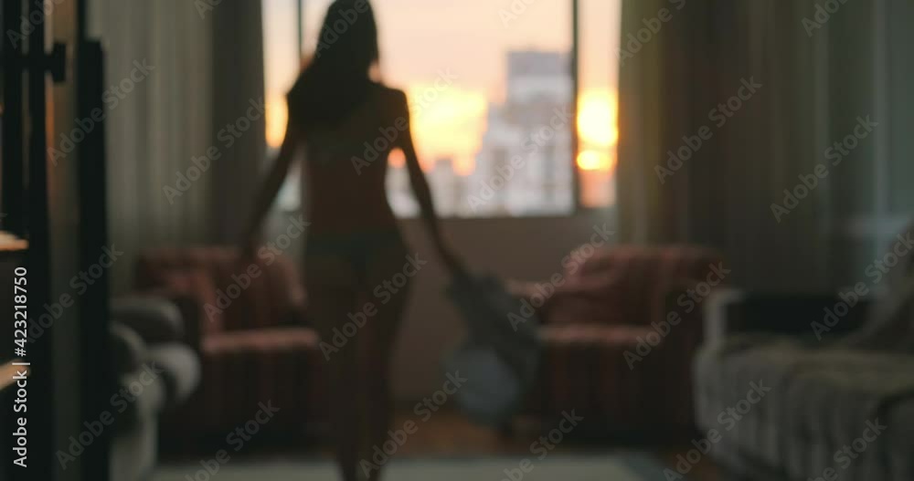 Young woman takes off her summer dress and dances near the wide window in the apartment at sunset