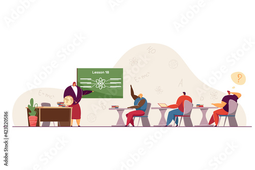 Students at physics lesson in classroom. Female teacher explaining and pointing at blackboard, pupils sitting at desks, black girl raising hand to answer. School, university, education concept