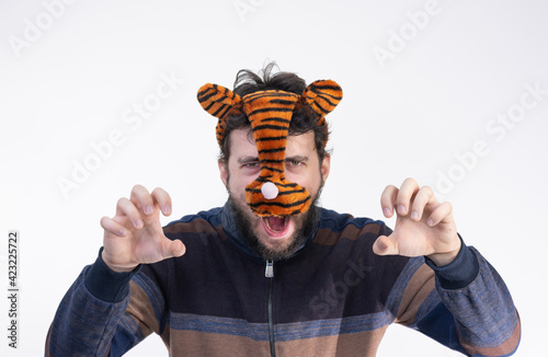 A young dark-haired man with a beard and a tiger mask poses as a tiger, on a light background with a bokeh effect