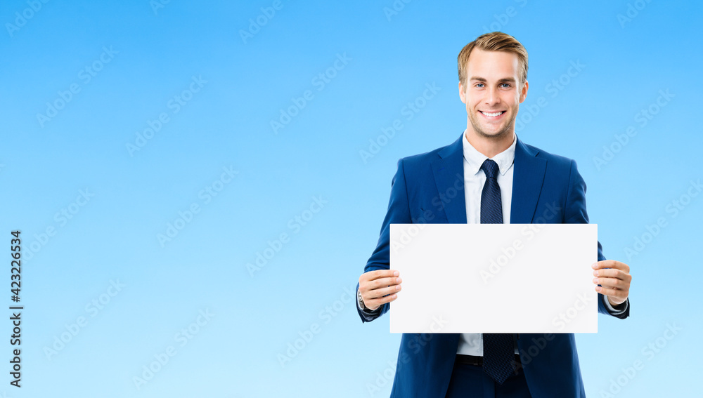 Businessman showing white paper signboard with copyspace area, over blue color background. Portrait of happy smiling confident man in business studio concept.