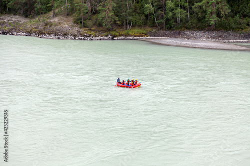 A team of people rafting in orange life jackets on an rubber boat of blue and yellow colors along a mountain river against the background of a rocky shore with a forest.