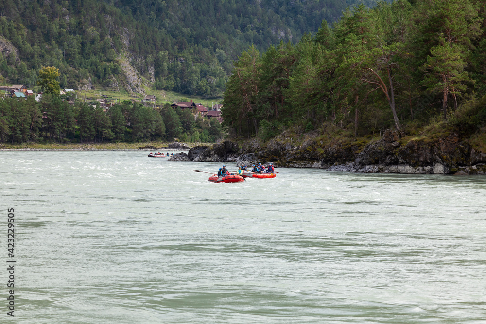 A team of people rafting in orange life jackets on an rubber boat of blue and yellow colors along a mountain river against the background of a rocky shore with a forest.