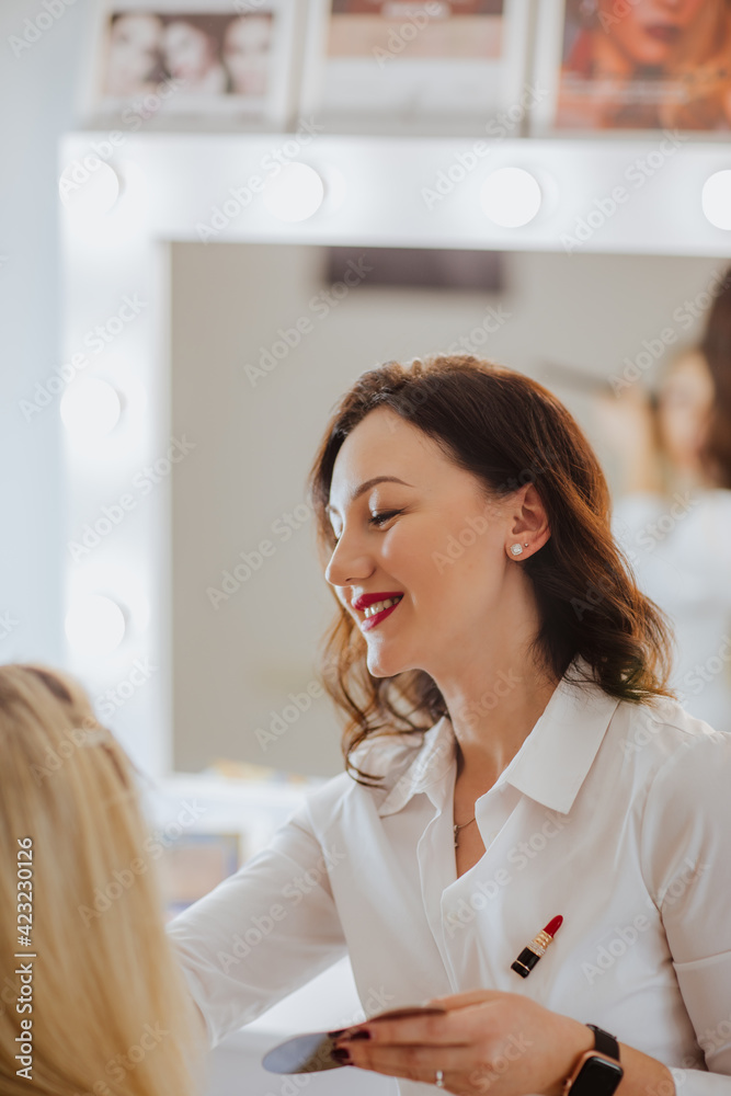 Pretty brunette woman in white shirt with red lips making make up for a blond model in her cabinet. Pretty brunette woman in white shirt with red lips making make up for a blond model in her cabinet.