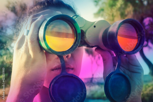 A girl looking at the horizon through a pair of military binoculars. Colorful prism reflection shades overlaid on her face. 