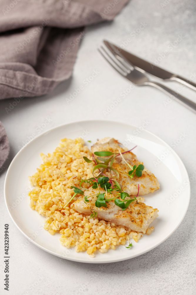 Baked fish hake or pollock and garnish bulgur and microzelenium in a plate on the table. Healthy food Baked fish hake or pollock and garnish bulgur and microzelenium in a plate on the table. Healthy food