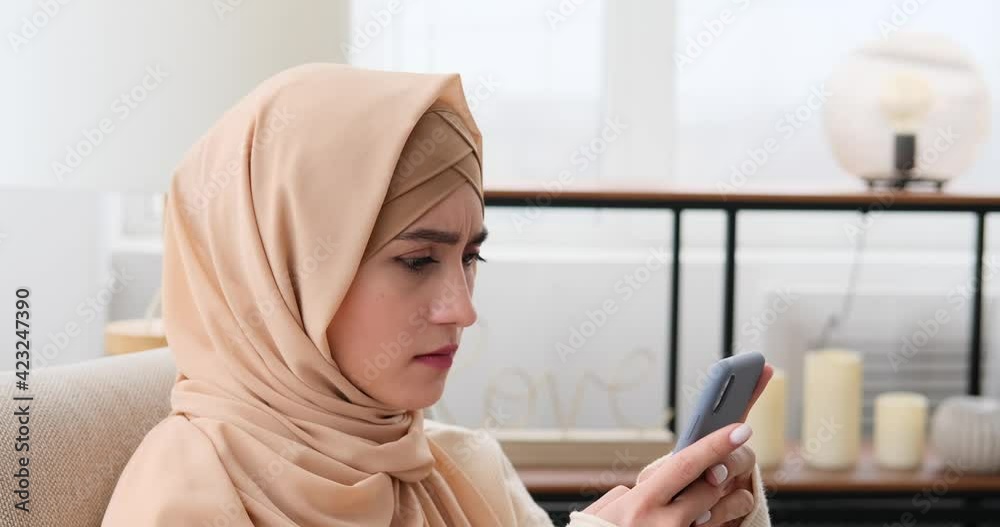 Displeased muslim woman in hijab text messaging using mobile phone at home