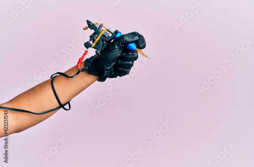 Hand of young hispanic man using tattoo machine over isolated pink background.