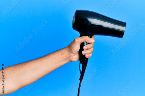 Hand of hispanic man holding hair dryer over isolated blue background.