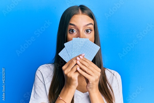 Young hispanic woman playing gambling poker covering face with cards afraid and shocked with surprise and amazed expression, fear and excited face.