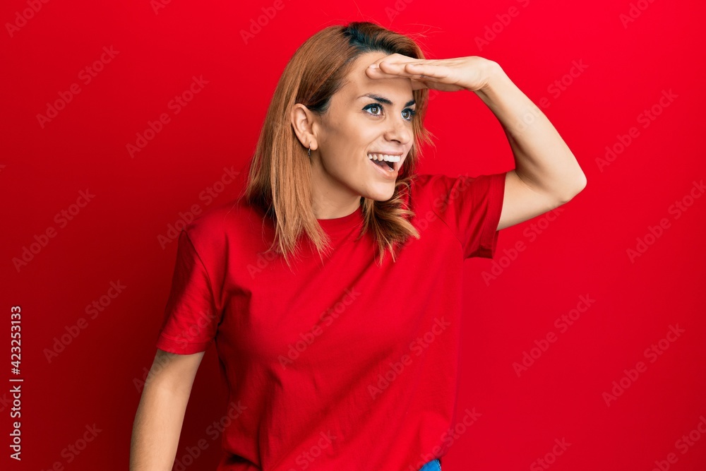 Hispanic young woman wearing casual red t shirt very happy and smiling looking far away with hand over head. searching concept. Hispanic young woman wearing casual red t shirt very happy and smiling looking far away with hand over head. searching concept.