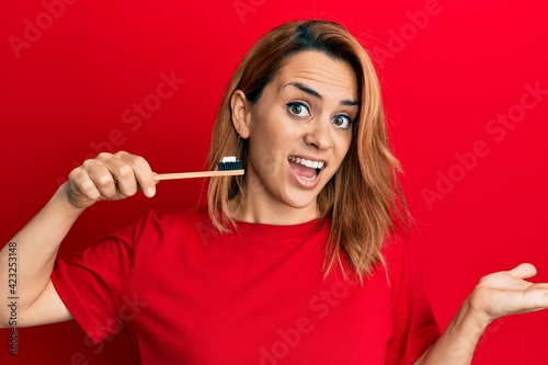 Hispanic young woman holding toothbrush with toothpaste celebrating achievement with happy smile and winner expression with raised hand