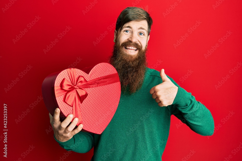 Redhead man with long beard holding valentine gift smiling happy and positive, thumb up doing excellent and approval sign Redhead man with long beard holding valentine gift smiling happy and positive, thumb up doing excellent and approval sign