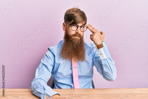 Obraz na plátne Young irish redhead man wearing business shirt and tie sitting on the table shooting and killing oneself pointing hand and fingers to head like gun, suicide gesture