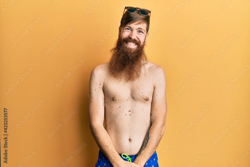 Redhead man with long beard wearing swimsuit and sunglasses looking positive and happy standing and smiling with a confident smile showing teeth