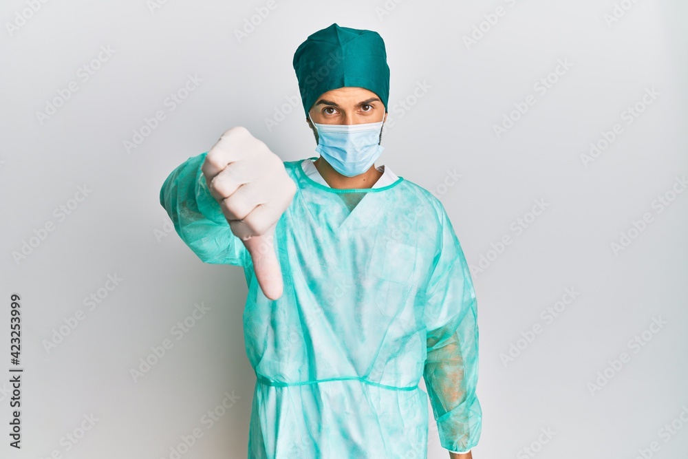 Young handsome man wearing surgeon uniform and medical mask looking unhappy and angry showing rejection and negative with thumbs down gesture. bad expression. Young handsome man wearing surgeon uniform and medical mask looking unhappy and angry showing rejection and negative with thumbs down gesture. bad expression.