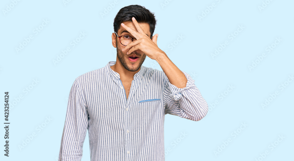Young handsome man wearing casual clothes and glasses peeking in shock covering face and eyes with hand, looking through fingers with embarrassed expression. Young handsome man wearing casual clothes and glasses peeking in shock covering face and eyes with hand, looking through fingers with embarrassed expression.