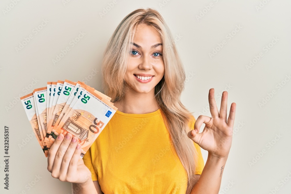 Beautiful caucasian blonde girl holding bunch of 50 euro banknotes doing ok sign with fingers, smiling friendly gesturing excellent symbol - obrazy, fototapety, plakaty 