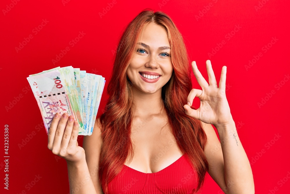 Young redhead woman holding hong kong dollars banknotes doing ok sign with fingers, smiling friendly gesturing excellent symbol Young redhead woman holding hong kong dollars banknotes doing ok sign with fingers, smiling friendly gesturing excellent symbol